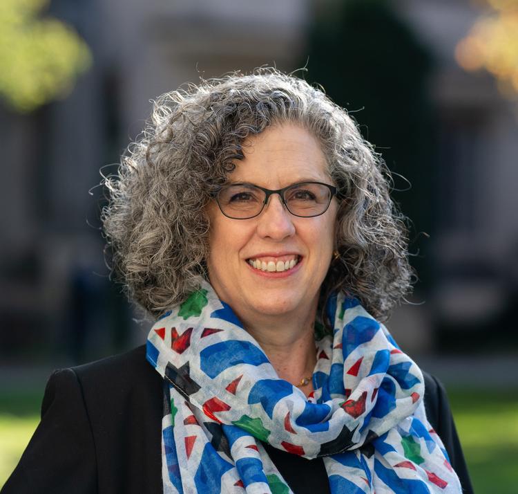 A photo of a woman with glasses and curly hair, wearing a colorful scarf, in an outdoor setting.